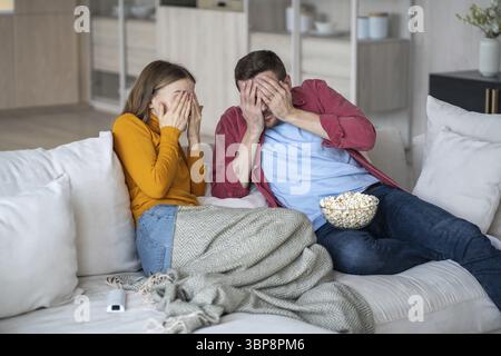 Homme choqué et femme fermant les visages avec les mains effrayées par le thriller terrifiant. Couple de famille effrayé regardant des séries de films ensemble assis sur le canapé Banque D'Images