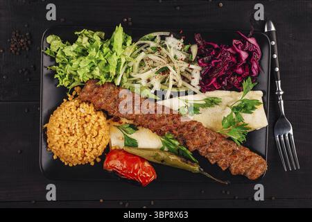 Nourriture, cuisine turque adana kebab avec légumes frais, boulgour et pain lavash sur assiette noire sur table en bois Banque D'Images