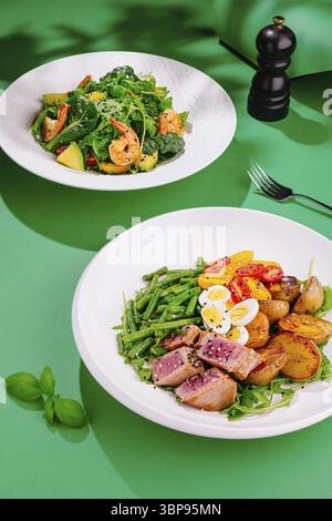 Nourriture, deux assiettes blanches avec de savoureuses salades fraîches avec des crevettes, thon, roquette, tomates et œufs sur fond vert Banque D'Images