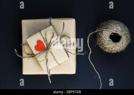Boîte-cadeau fabriquée à la main enveloppée de papier artisanal avec coeur en bois rouge, corde et noeud sur fond noir. Vue de dessus, plan d'appartement Banque D'Images
