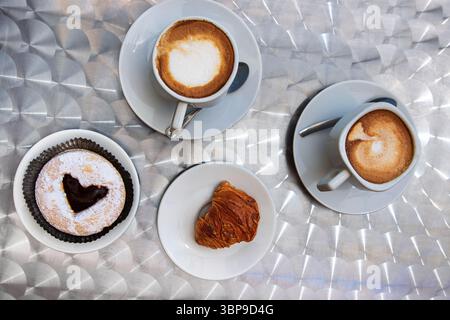 Vue aérienne du petit-déjeuner italien avec deux cappuccinos, une pâtisserie au chocolat et un sfogliatelle sur des plats blancs au sommet d'une table métallique texturée Banque D'Images
