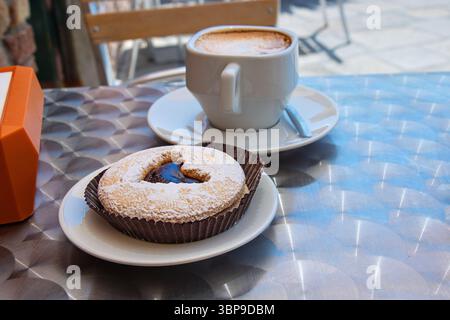 Une tasse fraîche de cappuccino dans un mug en céramique blanche servi avec une pâtisserie fourrée au chocolat surmontée d'un motif coeur, placée sur une table métallique dans un ou Banque D'Images