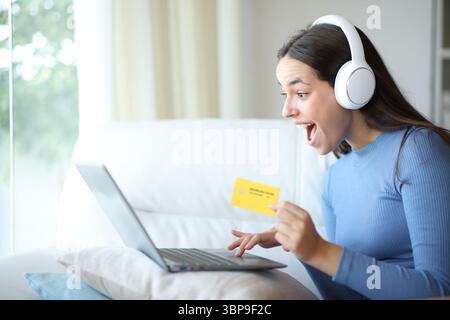 Femme étonnée achetant des médias en ligne en utilisant la carte de crédit et l'ordinateur portable assis sur un canapé dans un intérieur de maison Banque D'Images