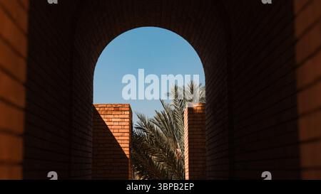 Vue sur une arche ensoleillée encadrée par des murs de briques, menant à des palmiers luxuriants à Babylone Irak. Mélange parfait d'histoire et de nature. Banque D'Images