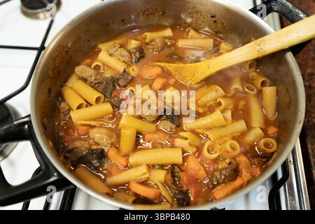 De délicieuses pâtes avec de la viande et des légumes sont cuites dans une casserole sur la cuisinière par un chef à l'aide d'une cuillère en bois Banque D'Images