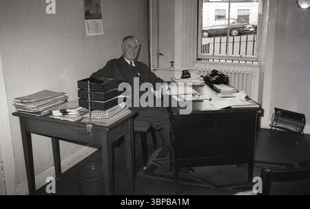 1967, historique, un homme mûr vêtu d'un costume rayé et d'une cravate, regarde la caméra, assis à son bureau situé près d'une fenêtre d'un petit bureau municipal d'une compagnie maritime, Édimbourg, Écosse, Royaume-Uni. Un tapis sous le bureau et un petit chauffage électrique au sol. Calendrier sur le mur derrière lui. Banque D'Images