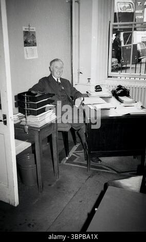 1967, historique, tous sourires, un homme mûr en costume à rayures et cravate, regarde la caméra, assis à son bureau situé près d'une fenêtre d'un petit bureau municipal d'une compagnie maritime, Édimbourg, Écosse, Royaume-Uni. Un petit tapis sous le bureau et bac en métal. Calendrier sur le mur derrière lui. Banque D'Images