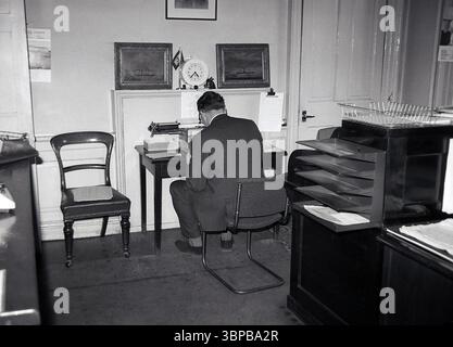 Années 1960, historique, à l'intérieur d'un bureau de ville, un gentleman mature en costume & cravate, assis à un petit bureau en bois, tentant de taper une lettre sur une machine à écrire manuelle, Édimbourg, Écosse, Royaume-Uni. Banque D'Images
