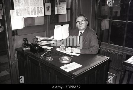Années 1960, historique, un cadre masculin en costume & cravate assis à un petit bureau en bois, dans les bureaux de la ville d'une compagnie maritime, Édimbourg, Écosse, Royaume-Uni. Un cendrier en métal, buvard et deux téléphones de l'époque sur le bureau. Banque D'Images