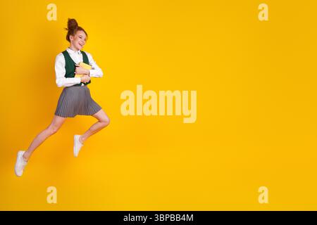 Écolière énergique sautant joyeusement dans un uniforme élégant tout en tenant des livres sur un fond jaune vif Banque D'Images