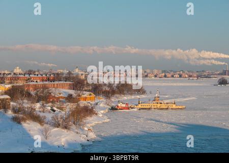 Vue hivernale de Suomenlinna vers Helsinki. Mer gelée, navires amarrés, bâtiments historiques et monuments au loin. Banque D'Images