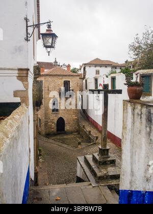 Croix médiévale en pierre et étroite rue pavée dans le centre historique de Óbidos, Portugal Banque D'Images