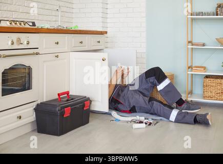 Plombier masculin en uniforme de travail couché sur le sol, fixant les tuyaux d'évier de cuisine, fournissant le service de plomberie. Banque D'Images