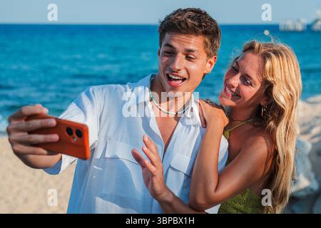 Un couple joyeux profitant de leur temps à la plage tout en prenant un délicieux selfie avec un smartphone. Ils capturent le moment rempli de Banque D'Images
