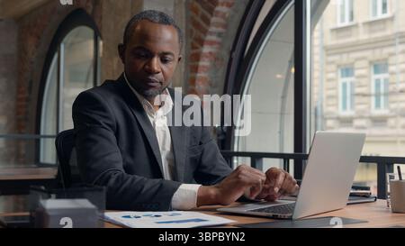 Entrepreneur afro-américain d'âge moyen dactylographiant ordinateur portable homme d'affaires bureau homme d'affaires homme masculin travaillant avec pc en ligne données de vérification d'emploi dans Banque D'Images