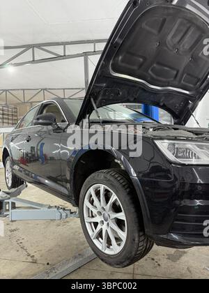 Cette image montre une voiture noire surélevée sur un pont élévateur hydraulique à l'intérieur d'un garage de service automobile. Le capot est ouvert, révélant une partie du compartiment moteur. Le Banque D'Images