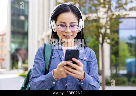 Une jeune femme asiatique portant des lunettes et des écouteurs sourit en regardant son smartphone à l'extérieur. Banque D'Images