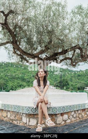 Belle jeune femme asiatique assise sous un grand olivier entouré d'un jardin tropical le week-end Banque D'Images