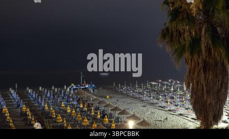 scène nocturne d'une plage de durres en albanie. des rangées de parasols fermés, certains illuminés, bordent le rivage sablonneux sous un ciel sombre, avec la mer calme i. Banque D'Images