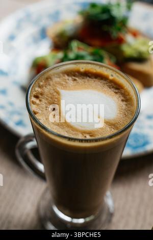 Une tasse de café avec un coeur dessiné sur le dessus est assise sur une table à côté d'une assiette de nourriture. Le café est brun et mousseux, et le cœur est blanc et légèrement Banque D'Images