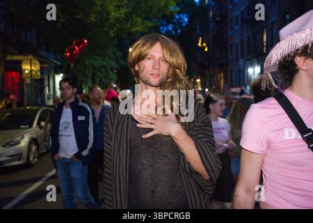 Une personne avec de longs cheveux blonds, portant une robe rayée sur un haut en maille, se tient à un événement de rue nocturne. D'autres personnes, dont une portant une épingle Banque D'Images