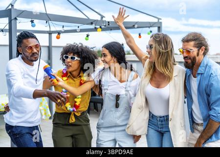 Groupe d'amis divers célébrant sur un toit, chantant karaoké, dansant joyeusement et profitant d'une fête d'été dynamique ensemble Banque D'Images