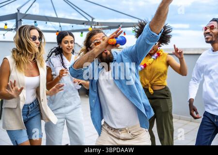 Groupe diversifié d'amis célébrant sur un toit dynamique, chantant et dansant joyeusement ensemble, embrassant l'esprit insouciant de l'été Banque D'Images