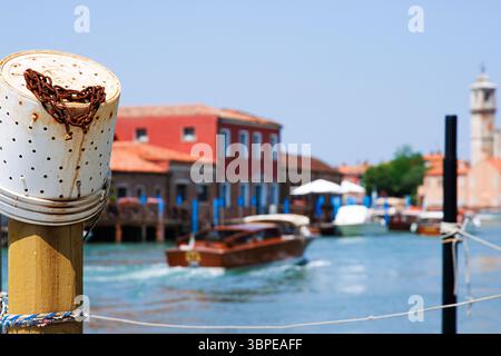 Murano, Province de Venise, Italie, 14 juin 2025. Poteau d'amarrage avec seau de pêche en plastique avec chaîne rouillée et un bateau à moteur se déplaçant le long du turquo Banque D'Images