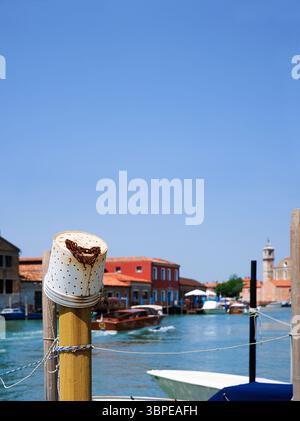 Murano, Province de Venise, Italie, 14 juin 2025. Poteau d'amarrage avec seau de pêche en plastique avec chaîne rouillée et un bateau à moteur se déplaçant le long du turquo Banque D'Images