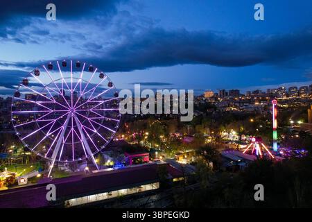 Une grande roue brillante avec des lumières violettes au crépuscule dans Victory Park, Erevan, Arménie, surplombant l'horizon de la ville et les attractions du parc d'attractions. Banque D'Images