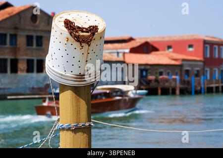 Poste d'amarrage avec seau de pêche en plastique avec chaîne rouillée et un bateau à moteur se déplaçant le long de l'eau du canal turquoise passant devant des bâtiments colorés à Murano Banque D'Images