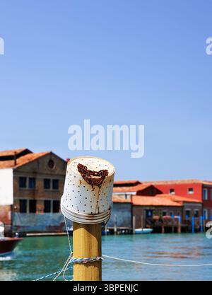 Poste d'amarrage avec seau de pêche en plastique avec chaîne rouillée et un bateau à moteur se déplaçant le long de l'eau du canal turquoise passant devant des bâtiments colorés à Murano Banque D'Images