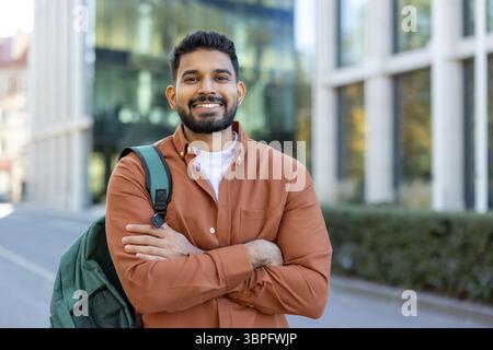 Un étudiant indien souriant avec un sac à dos se tient confiant devant un bâtiment moderne, les bras croisés Banque D'Images