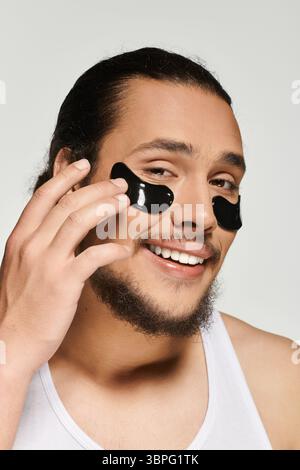 Jeune homme barbu sourit tout en appliquant sous les patchs oculaires pour un regard frais pendant les soins personnels. Banque D'Images
