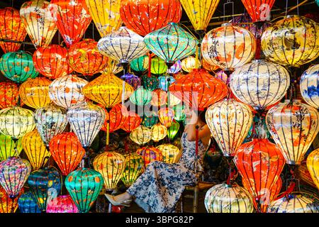 Da Nang, Vietnam-18 mars 2025 : femme en robe florale est assis parmi les lanternes de soie brillantes, immergé dans un monde éblouissant de couleur, de lumière et de charme culturel Banque D'Images