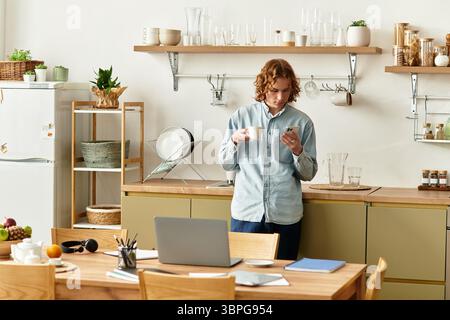 Jeune homme se tient dans une cuisine élégante, sirotant du café tout en regardant son téléphone. Banque D'Images