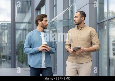 Deux hommes d'affaires conversant à l'extérieur près d'un bâtiment moderne en verre, l'un tenant une tasse de café, l'autre une tablette. Banque D'Images