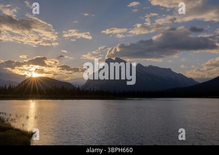 Vue du soleil doré éclatant à travers les nuages sur le lac tranquille et les montagnes silhouettes, reflétant le ciel serein, Vermilion Lake, Alberta, Canada. Banque D'Images