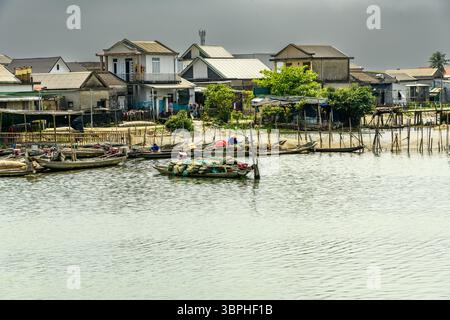 Une scène tranquille de maisons vietnamiennes traditionnelles construites sur pilotis le long d'une rivière calme. Plusieurs petits bateaux de pêche sont amarrés le long des maisons, wit Banque D'Images