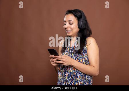 Jeune belle femme avec des écouteurs sourit tout en parcourant la musique sur smartphone. Jolie mannequin indienne appréciant ses morceaux préférés via un casque sans fil pendant qu'elle envoie des SMS sur un appareil mobile. Banque D'Images