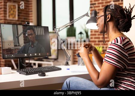 Femme noire souriante assise au bureau, participe à une réunion virtuelle avec un client potentiel. Employée afro-américaine travaillant à domicile, ayant une discussion en ligne avec le manager masculin sur l'appel vidéo. Banque D'Images