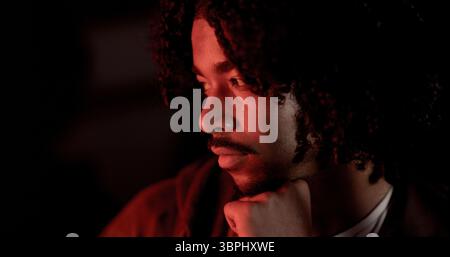 Gros plan portrait d'un jeune homme réfléchi avec des cheveux bouclés et une barbe, illuminé par une lumière rouge dramatique, créant un mystère et contemplatif Banque D'Images