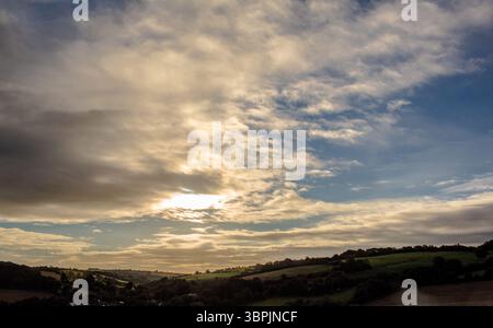 La lumière du soleil filtre à travers le ciel spectaculaire au-dessus des collines verdoyantes et des terres agricoles dans un design plat Banque D'Images