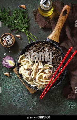 Nouilles épicées, cuisine traditionnelle coréenne. Nouilles de soba de sarrasin et calmars avec sauce à la crème aigre sur une table en pierre sombre. Vue de dessus de l'arrière-plan de la mise à plat Banque D'Images