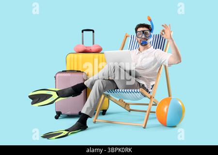 Jeune homme d'affaires avec ordinateur portable montrant OK dans la chaise longue et accessoires de plage sur fond bleu. Concept de vacances d'été Banque D'Images