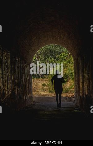 La silhouette d'un coureur isolé émerge d'un tunnel sombre, encadré par une végétation luxuriante à l'extérieur. La scène capture l'essence de la solitude et de la détermination dans Banque D'Images