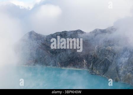 Vue sur les eaux turquoise éthérées du lac rencontrer le cratère accidenté et fumé du volcan, enveloppé dans la brume, une scène de beauté naturelle brute, cratère du volcan Ijen, EA Banque D'Images