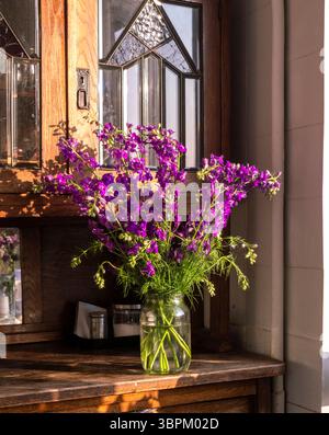 Un groupe de fleurs violettes éclatantes remplit un vase en verre transparent reposant sur une table en bois. La lumière du soleil du matin traverse une grande fenêtre, illuminant Banque D'Images
