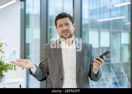 Portrait d'un jeune homme d'affaires inquiet debout dans un costume dans le bureau, écartant les bras et regardant la caméra tristement. Banque D'Images