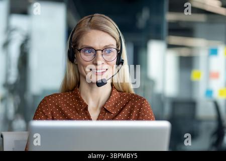Une femme blonde souriante portant un casque et des lunettes travaille sur un ordinateur portable dans un cadre de bureau moderne, éventuellement au service à la clientèle. Banque D'Images